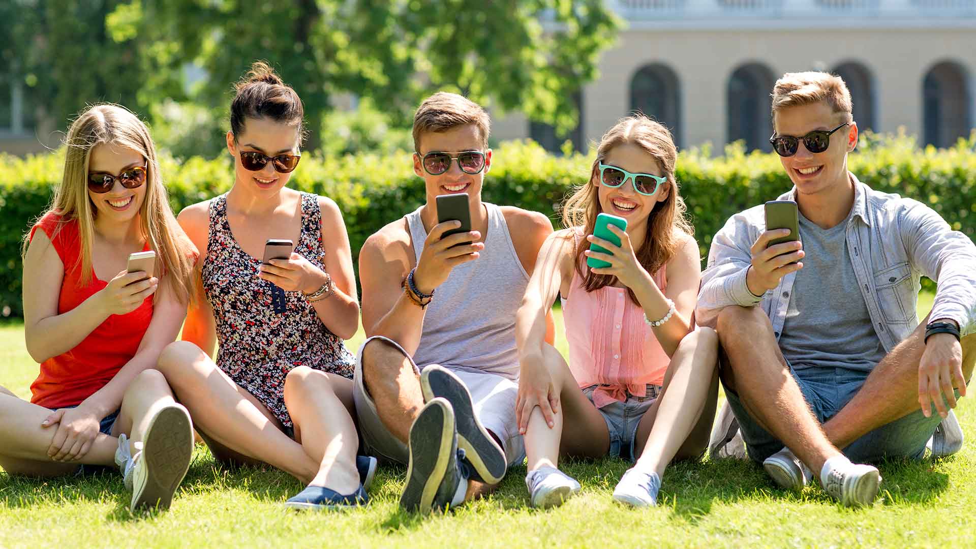 five students sitting on grass looking at their phones with sunglasses on
